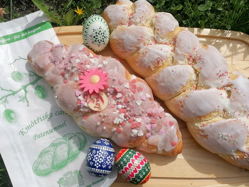 Zwei verzopfte Patensemmeln mit Zuckerguss und bunten Streuseln liegen auf einem Holzbrett. Daneben sind drei verzierte Ostereier und eine Bäcker-Papiertüte zu sehen. Sorbische_Ostereier_und_Patenzöpfe_zu_Gründonnerstag