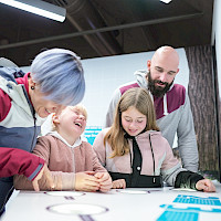 Eine Familie mit zwei Kindern interagiert mit einer interaktiven Ausstellung. Die Kinder lachen und zeigen auf etwas auf einem Tisch, während die Eltern zuschauen.