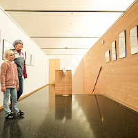 Eine Frau und ein Kind stehen in einem Ausstellungsraum und betrachten Kunstwerke. Die Wände sind mit Bildern und Holzverkleidungen geschmückt.