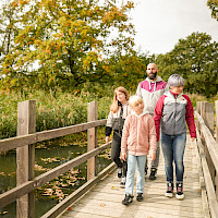 ine Familie spaziert über eine Holzbrücke in einer herbstlichen Landschaft. Die Bäume haben bunte Blätter und das Wasser unter der Brücke ist mit Laub bedeckt.