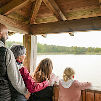Eine Familie steht in einem überdachten Aussichtsturm aus Holz und blickt auf einen See. Ein Mann umarmt eine Frau, während zwei Mädchen neben ihnen stehen.