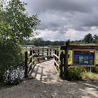 Ein Holzsteg führt zu einem See, daneben steht ein Informationstafel. Der Himmel ist bewölkt.