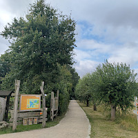 Ein Kiesweg führt durch eine grüne Landschaft mit Bäumen und einem Informationsschild. Der Himmel ist bewölkt mit einigen blauen Flecken.