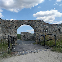 Das Bild zeigt eine steinerne Torbogenruine mit einem hölzernen Steg und Zaun davor. Im Hintergrund sind weitere Ruinen und Bäume unter einem blauen Himmel mit Wolken zu sehen.