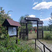 Ein Holz-Torweg steht in einem parkähnlichen Gelände, durch das man einen roten Torbogen sieht. Links daneben befindet sich eine Informationstafel.