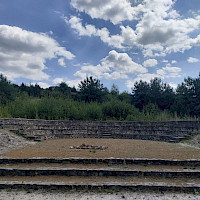 Das Amphitheater_Eine Steinterrasse mit Sitzreihen und einer kleinen Feuerstelle im Freien unter einem blauen Himmel mit Wolken. Dahinter liegt ein dichter Baumbestand.