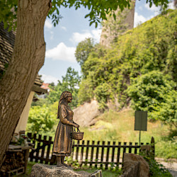 Eine Bronzestatue einer Frau mit einem Korb steht auf einem Stein im grünen Garten unterhalb eines Baumes. Im Hintergrund ist der Turm die Alte Wasserkunst zu sehen.  Im Vordergrund und im Hintergrund gibt es viel Grün und einen Holzzaun.