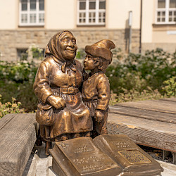 Eine Bronzestatue einer lachenden Großmutter und eines Kindes sitzt auf einer Holzbank mit einem offenen Buch davor. Hinter ihnen steht ein Gebäude.