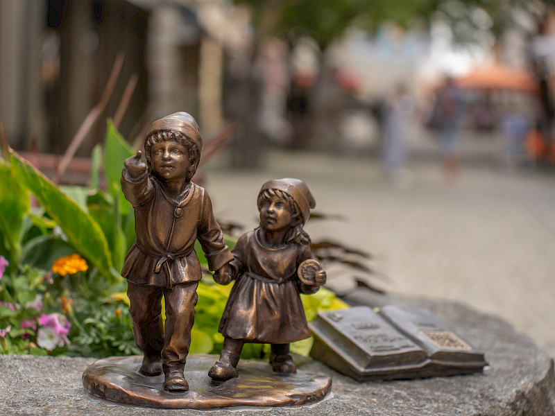 Zwei kleine Bronze-Figuren von Kindern stehen auf einem Stein. Neben ihnen liegt eine bronzene aufgeschlagene Buchskulptur. Im Hintergrund ist eine unscharfe Straßenszene zu sehen.