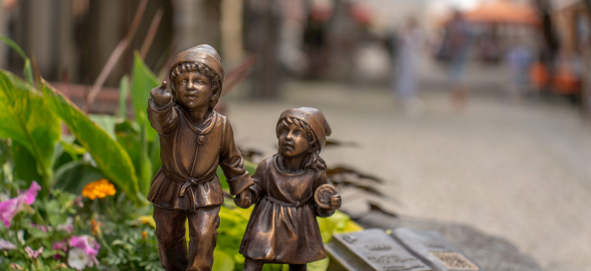 Zwei kleine Bronze-Figuren von Kindern stehen auf einem Stein. Neben ihnen liegt eine bronzene aufgeschlagene Buchskulptur. Im Hintergrund ist eine unscharfe Straßenszene zu sehen.