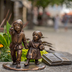 Zwei kleine Bronze-Figuren von Kindern stehen auf einem Stein. Neben ihnen liegt eine bronzene aufgeschlagene Buchskulptur. Im Hintergrund ist eine unscharfe Straßenszene zu sehen.