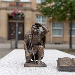 Ein kleines Bronzedenkmal eines knienden Mannes (Riese Sprejnik) mit einem Speer in der Hand steht auf einer Steinoberfläche. Daneben liegt ein weiteres Bronzedenkmal in Form eines offenen Buches. Im Hintergrund ist ein Gebäude mit Fenstern zu sehen.