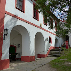 Ein weißes Gebäude mit roten Akzenten und Bögen ist auf der linken Seite zu sehen, während rechts eine Treppe zu einer Tür führt. Ein Schild mit der Aufschrift