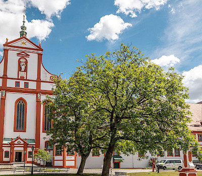 Klosterkirche und Klostergebäude_Ein imposantes Barockgebäude mit roten Akzenten, umgeben von grünen Bäumen und Rasenflächen unter einem blauen Himmel mit Wolken. Rechts daneben befindet sich ein weiteres Gebäude mit rotem Ziegeldach.