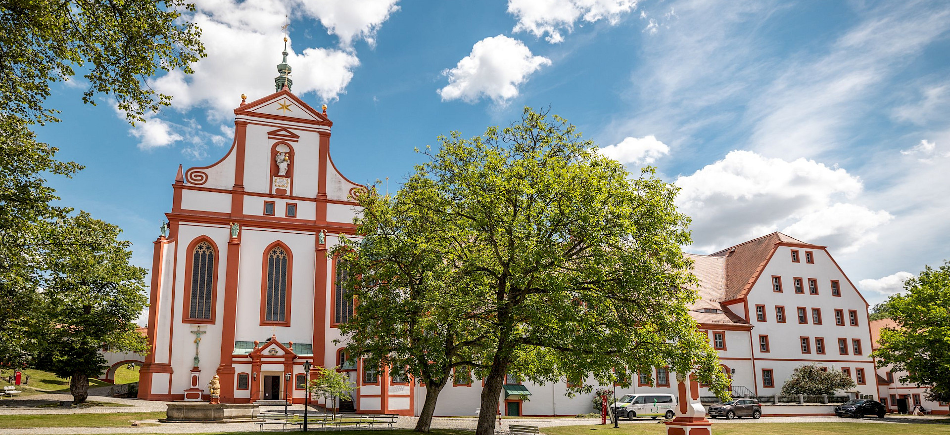 Klosterkirche und Klostergebäude_Ein imposantes Barockgebäude mit roten Akzenten, umgeben von grünen Bäumen und Rasenflächen unter einem blauen Himmel mit Wolken. Rechts daneben befindet sich ein weiteres Gebäude mit rotem Ziegeldach.