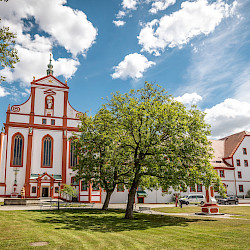Klosterkirche und Klostergebäude_Ein imposantes Barockgebäude mit roten Akzenten, umgeben von grünen Bäumen und Rasenflächen unter einem blauen Himmel mit Wolken. Rechts daneben befindet sich ein weiteres Gebäude mit rotem Ziegeldach.